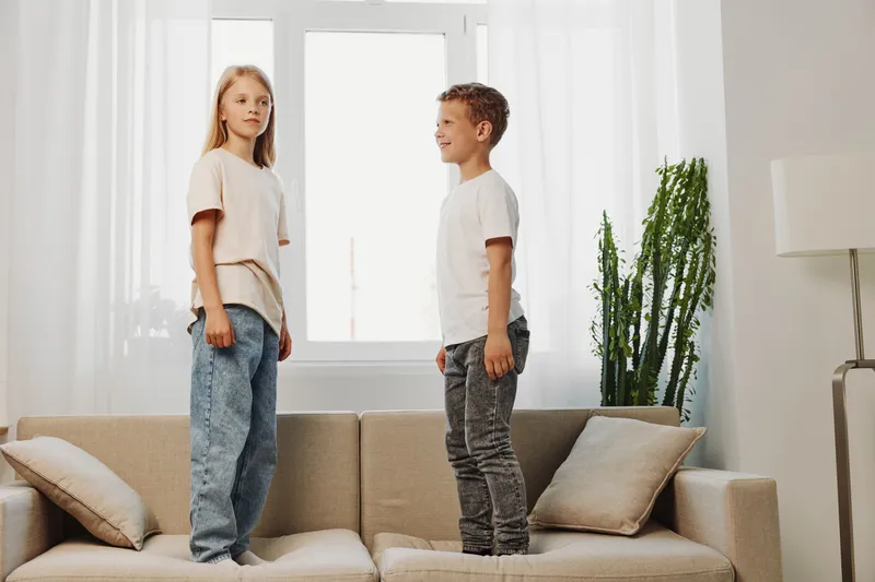 Two children standing on a sofa facing each other in a bright living room with natural light and minimalist decor, including green plants and cushions.