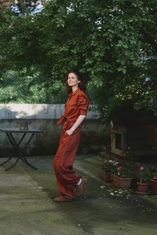 Woman outdoor lifestyle in orange jumpsuit walking through a garden, casual pose, bright greenery, and warm light. Natural, relaxed mood, everyday serenity and freshness