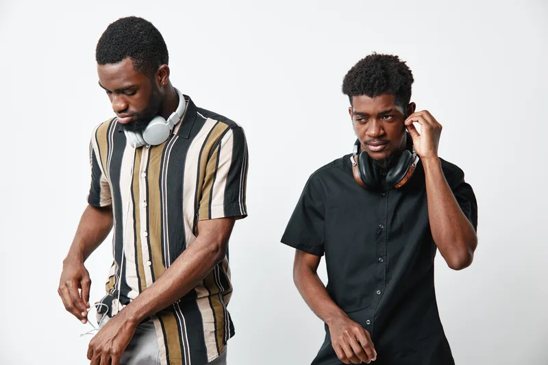 Two young black men with dark skin wearing headphones and casual clothes standing against white background. One man in striped shirt, the other in black shirt adjusting headphones.