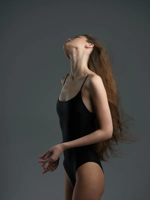 graceful woman in black bodysuit with long hair posing against gray background in studio