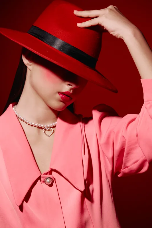 Fashionable woman in pink coat posing with red wide brim hat partially covering her face, wearing pearl necklace and heart pendant on red background. Concept of beauty, fashion, creative style.