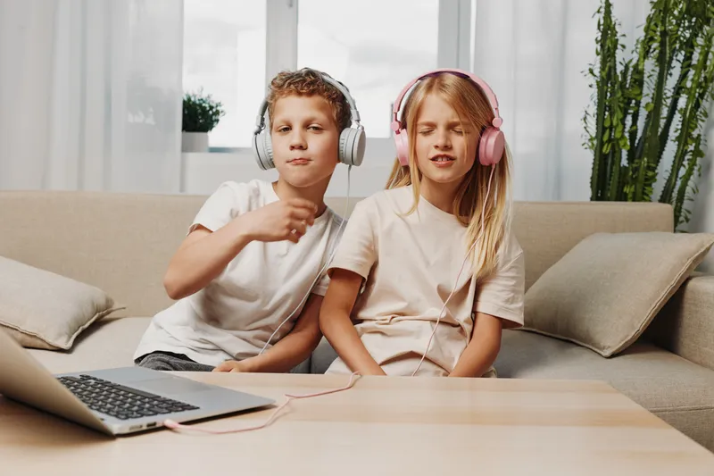 Two children with headphones enjoying music together on sofa in bright living room, sharing a moment of happiness and connection.