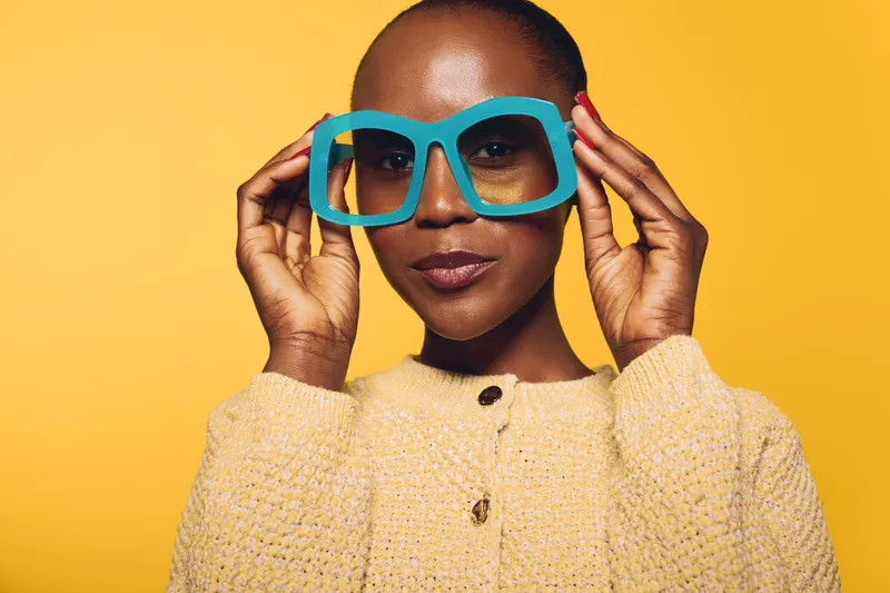 Confident black woman with dark skin wearing oversized blue glasses and a yellow sweater poses against a bright yellow background. Concept for fashion, style, and self-expression.
