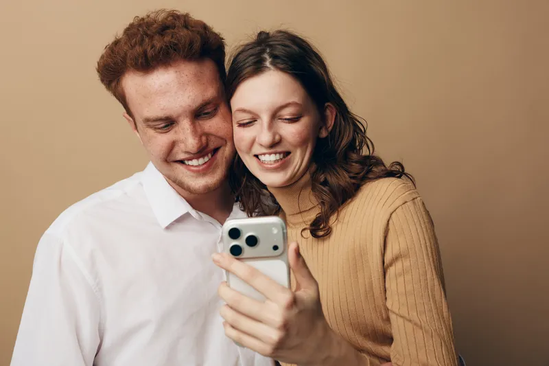 Smiling young couple with red hair and brown hair looking at smartphone together. They wear casual clothes and share a happy moment in front of beige background. Concept of friendship and technology.