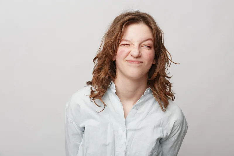 Young woman with curly hair wearing white shirt makes funny face with eyes closed and scrunched nose. Studio portrait on light background. Concept of humor, emotion, and casual style.