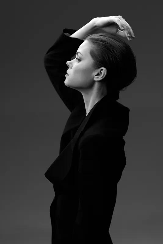Black and white portrait of a thoughtful woman in elegant dark clothing with hand raised against a dark background.