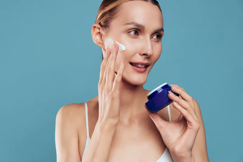 Woman apply face cream, holding cosmetic jar, fresh beauty skincare routine, moisturizer on skin, clean close up portrait, turquoise background, spa mood