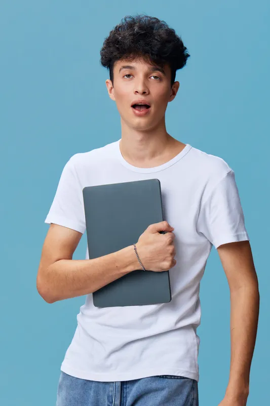 Young man holding laptop with surprised expression, wearing casual white t-shirt and jeans, standing against plain light blue background.