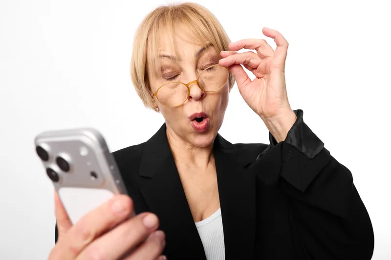 Woman with glasses surprised while holding a smartphone, adjusting eyewear in a business office portrait, blond hair, communication gesture, isolated white background, studio