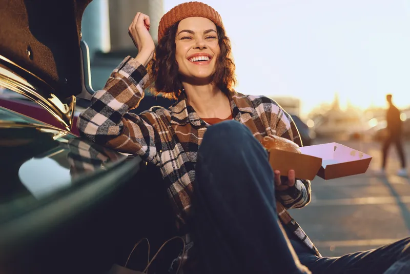 Woman with pizza sitting by car on street at sunset, beanie and plaid jacket, smile and relaxed vibe, capturing authenticity, candid lifestyle, golden hour glow, mindful living and emotional