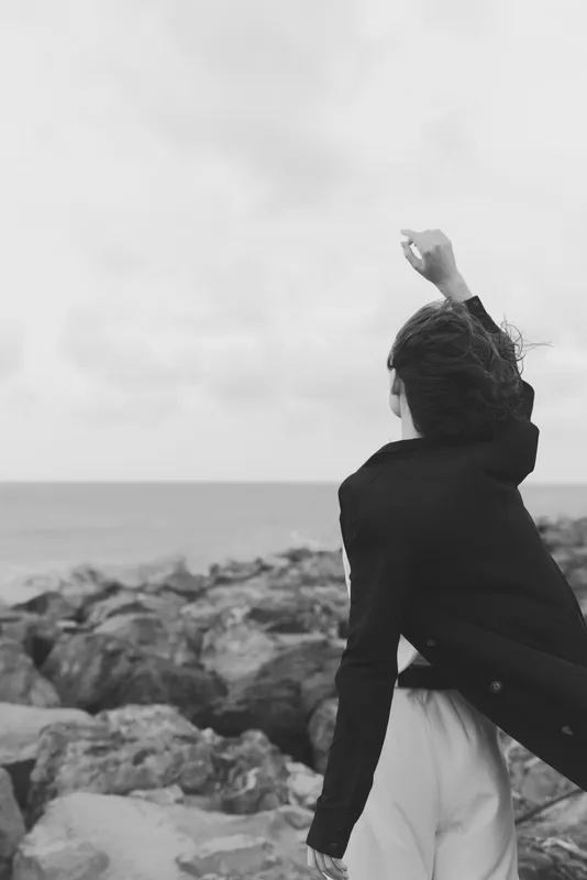 Woman stands on a rocky coast with wind in her hair, jacket fluttering, raising a fist toward the horizon, evoking freedom, resilience and bold outdoor spirit by the sea