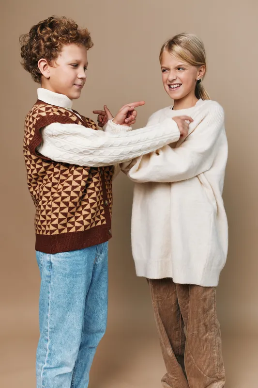 Playful children in cozy sweaters share a light moment, pointing at each other with smiles, capturing warmth, friendship, and casual style in a friendly indoor scene.