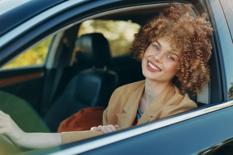 A smiling woman sits in a car seat, sunlit interior highlights her curly hair and blazer, conveying cheerful, casual lifestyle and confident independence on the road.