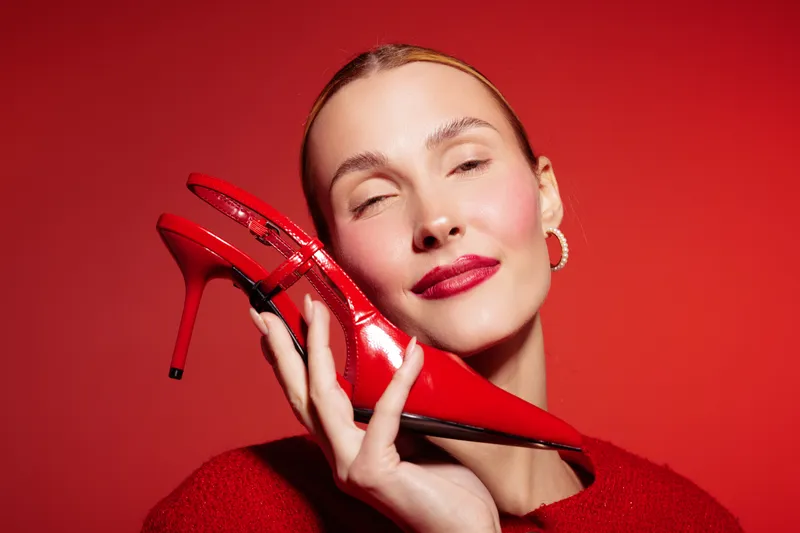 Woman with red lipstick holds shiny red high heel shoe near face on red background. Concept of fashion, beauty, style, and glamour with vibrant colors and confident expression.