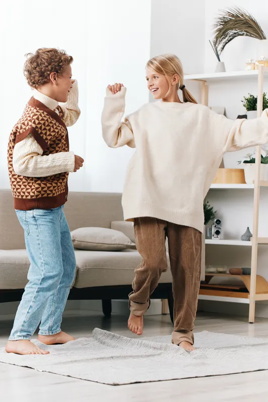 Two children dance joyfully indoors, wearing cozy sweaters and relaxed pants, sharing a playful moment in a bright living room with soft textures and warm natural light.
