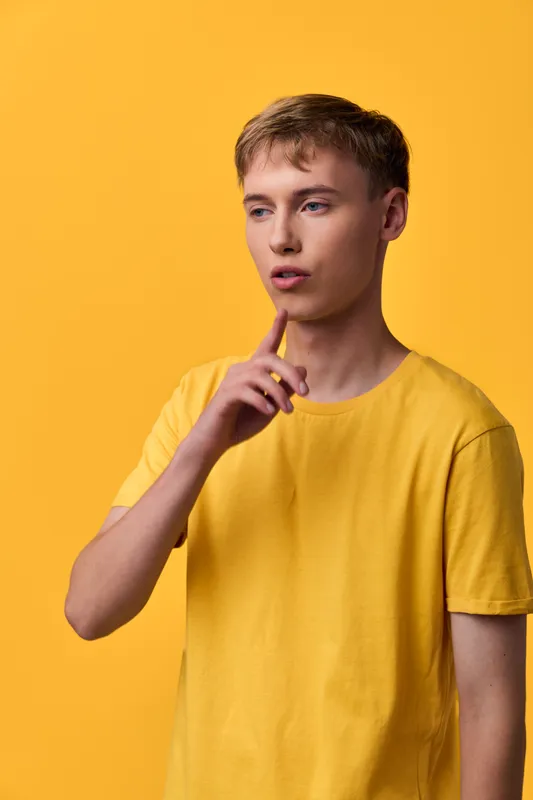 Thinking young man in a yellow shirt against a bright yellow backdrop, showcasing contemplation, focus, and curiosity with a thoughtful finger gesture