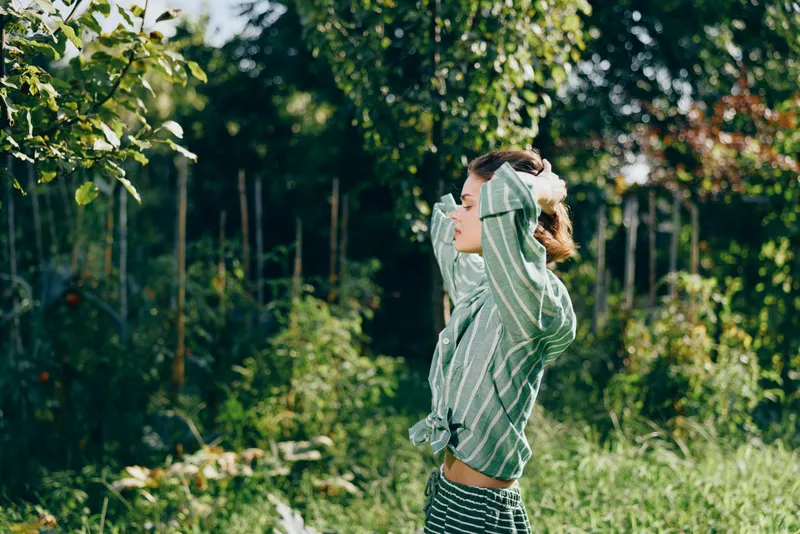 A woman outdoors in a sunlit garden stretches her arms above her head, enjoying fresh air and nature, a carefree, relaxed moment filled with light and joy.