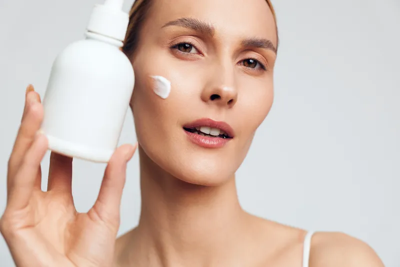 A young woman with clear skin holds a white bottle and applies cream on her cheek. The close-up shows skincare routine and natural beauty care on a light background.
