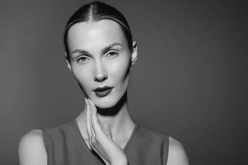 Portrait of a fair skinned white woman with slicked hair touching her neck against grey background.