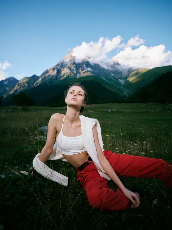 Outdoor athletic landscape with meadow and alpine mountains, an athletic pose in light top and red pants, sunny breeze and nature energy