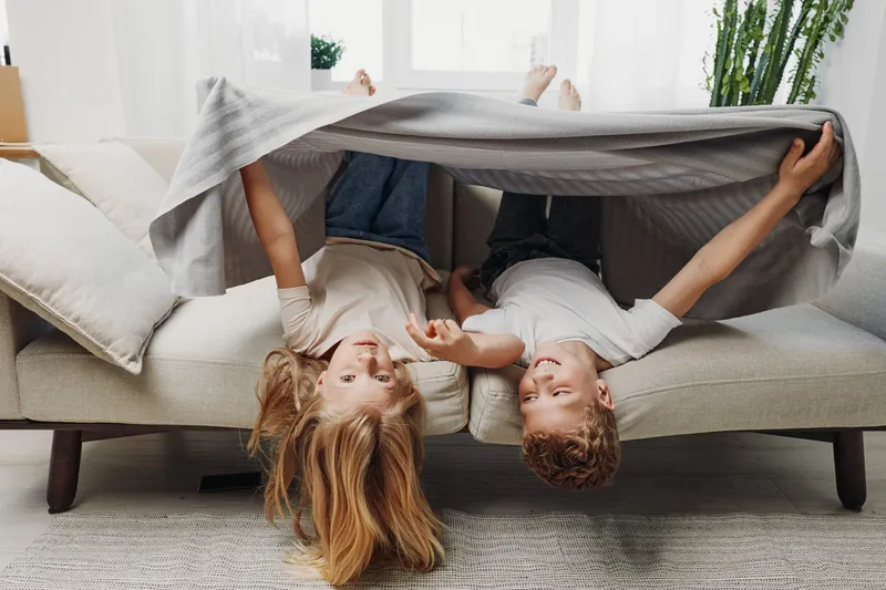 Two children lie upside down on a sofa covered by a blanket, smiling and playing in a bright living room with modern decor and plants.