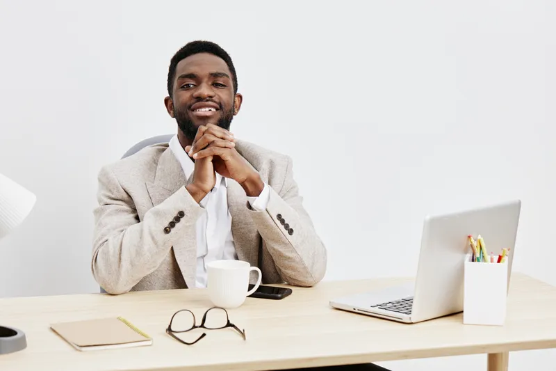 Smiling man sitting at a desk in an office environment, wearing a beige blazer and white shirt, looking confidently at the camera. Modern workspace setup with a laptop, notebook, and coffee cup, conve