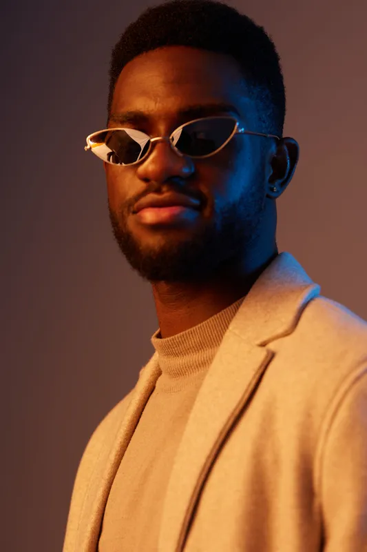 Stylish young man in sunglasses with a confident expression, wearing a beige blazer and sweater against a vibrant blue and orange background, embodying modern fashion and self assurance