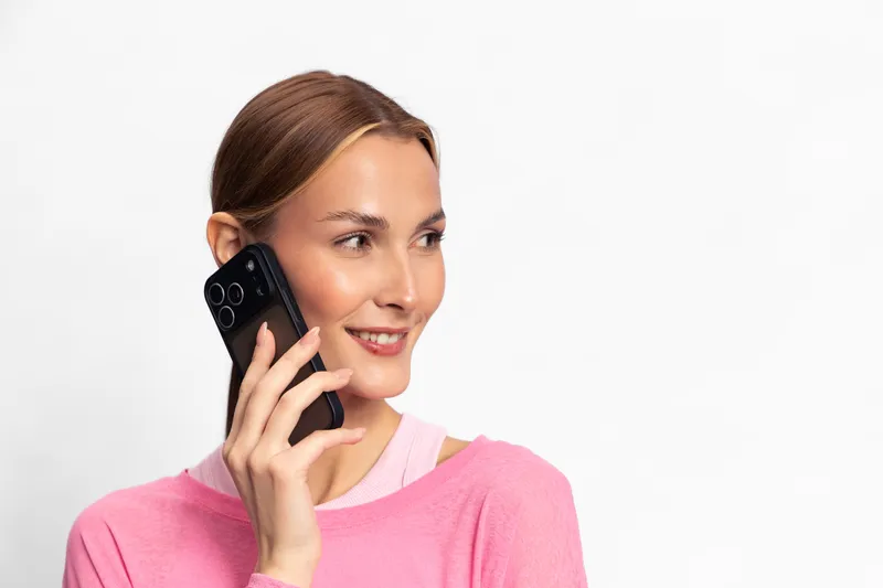 Smiling woman in pink sweater holds smartphone near ear, looking sideways. Bright studio background and casual outfit create a positive communication and technology concept.