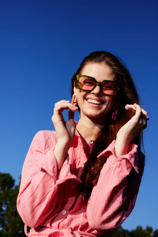 A cheerful young woman wearing sunglasses and a pink shirt, enjoying a sunny day outdoors, surrounded by greenery and a clear blue sky, radiating joy and positivity