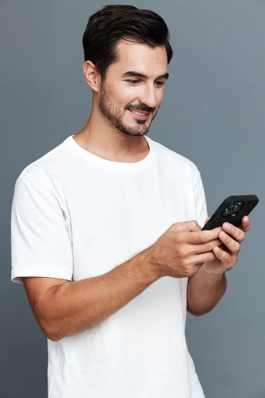 Full body smiling cheerful young man wearing a casual white t-shirt looks at his smartphone, engaged in conversation, against a solid grey background.