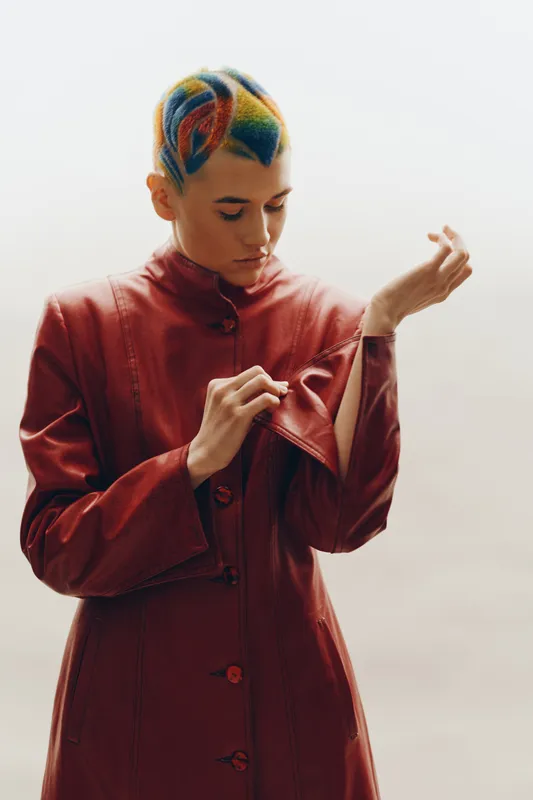 young woman with colorful short hair wearing a red leather jacket unzipping sleeve with focused expression on neutral background