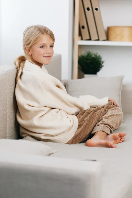 Young girl sits comfortably on a soft sofa wearing a cream knit sweater and cozy pants, creating a serene, candid portrait with warm natural light and relaxed posture.