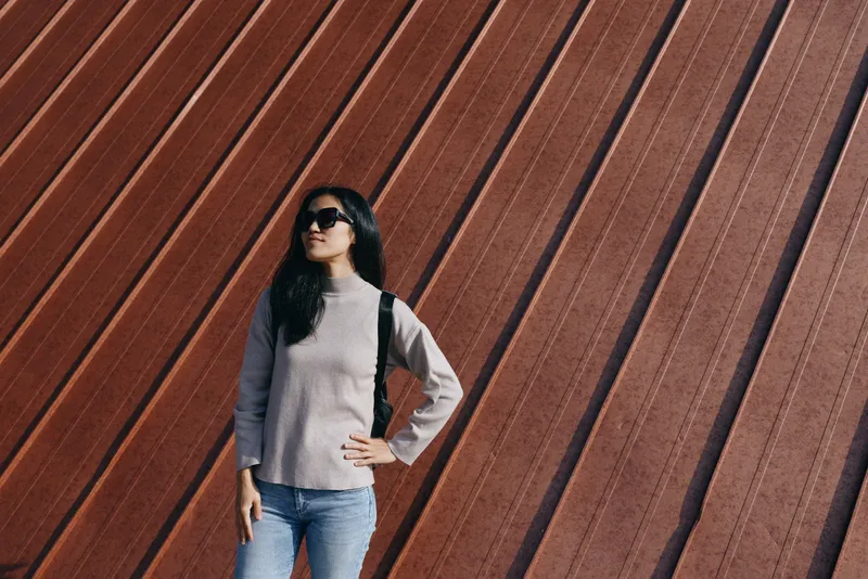 Woman in casual outfit stands beside a diagonal rust wall, sunglasses on, with a confident pose highlighting urban fashion, outdoor light, and textured architectural backdrop.