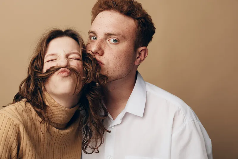 Young couple in casual clothes share a playful moment, man kissing woman on cheek while she makes a funny face with hair mustache. Concept of love, fun, and relationship joy.