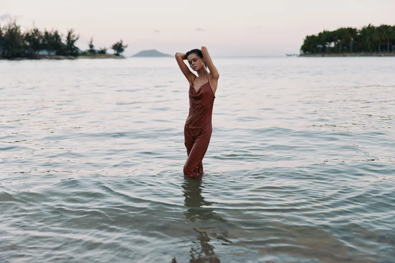 A solitary woman stands waist-deep in calm sea water, wearing a rust brown dress, arms raised, as soft sunset light creates a tranquil, reflective mood.