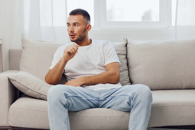 Thoughtful man sitting on a sofa, wearing a casual outfit while pondering ideas in a bright, modern living space. Ideal for lifestyle and mental health themes.