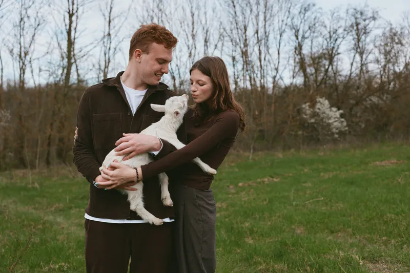 Smiling couple in casual clothing gently cradle a goat kid in a grassy park, warm sunlight softens the moment, conveying lifestyle, affection, and a love of nature.
