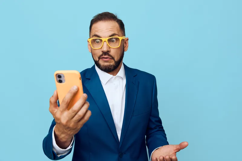 A professional man in a blue suit stands against a solid colored backdrop, expressing curiosity and surprise while capturing a selfie on his phone.