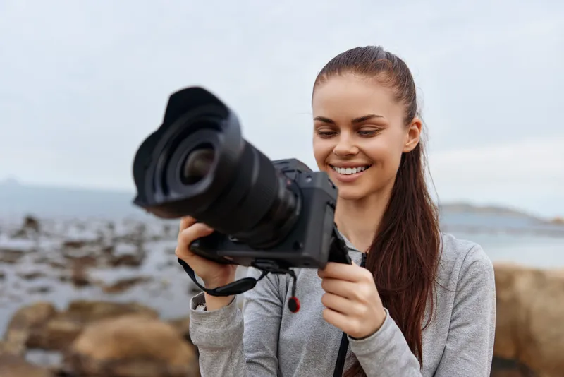 Young woman with long hair joyfully taking photos with a professional camera by the seaside, wearing a casual outfit in a serene outdoor setting