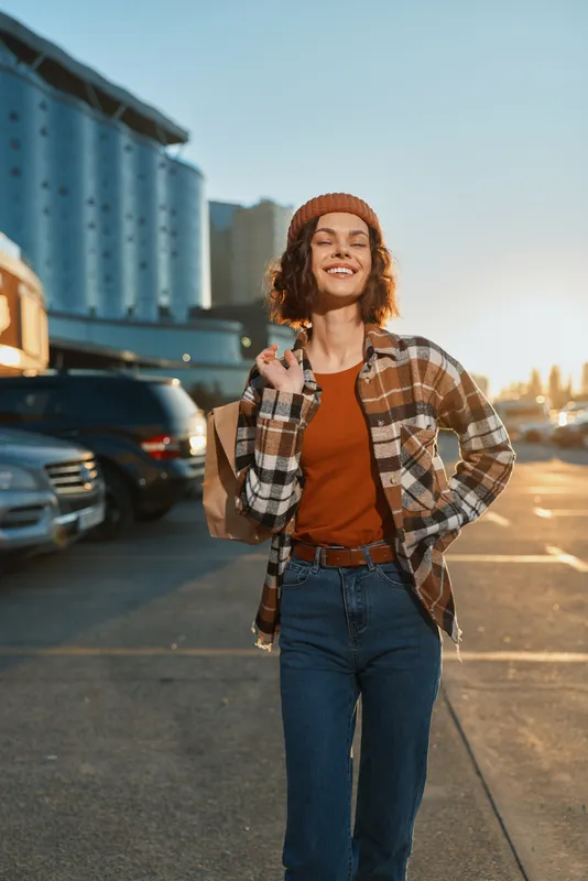 Woman smiling and walking in urban parking lot with shopping bag, sunlit golden hour glow, candid lifestyle portrait conveying authenticity, mindful living and emotional storytelling.