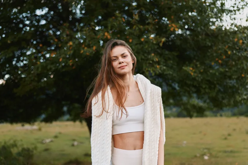 Young woman with long hair wearing white knitwear standing outdoors in nature with trees and grass in the background. Concept of lifestyle, relaxation, and casual fashion.