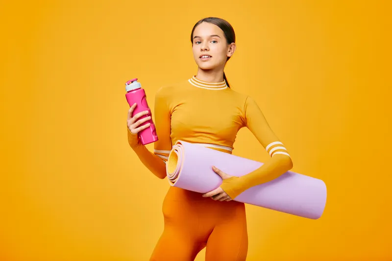 Woman in bright orange athletic wear holding a pink water bottle and a rolled purple yoga mat against a bold orange backdrop, conveying a dynamic fitness lifestyle and training focus.