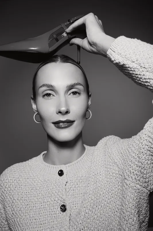 Caucasian woman with fair skin holds red high heel shoe above head wearing knitted sweater and hoop earrings.