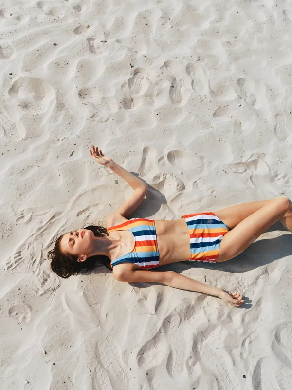 Sunlit woman lying on white sandy beach, wearing a bright striped bikini. Relaxed pose, arms at sides, carefree expression, capturing warm summer light and seaside calm, photography vibe.