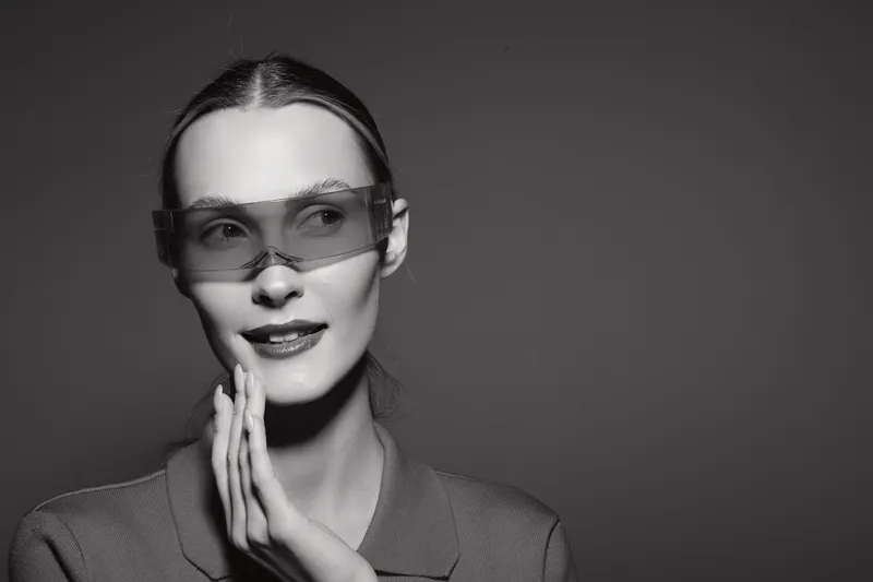 Portrait of a young white woman with pale skin wearing modern transparent sunglasses and a textured jacket in a studio with dark background.