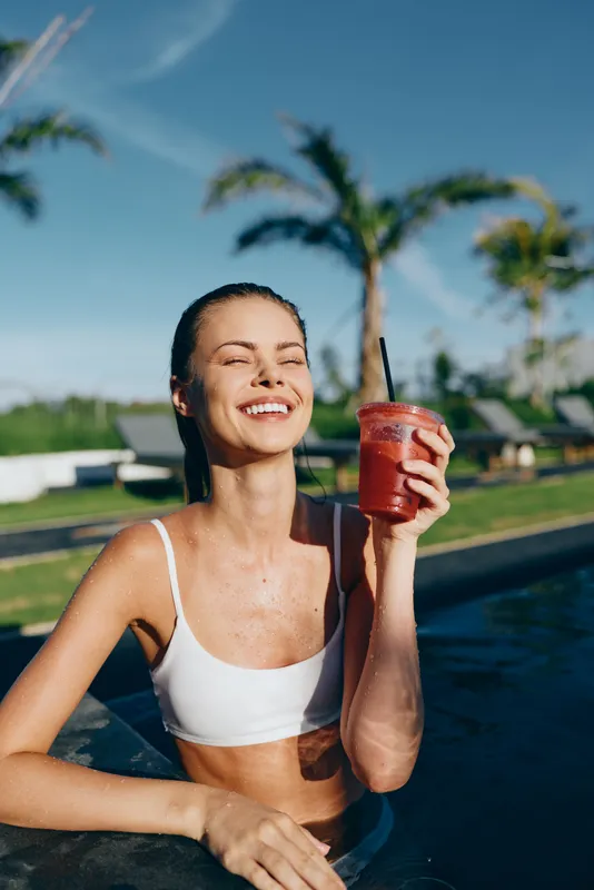Smiling woman in white bikini holding a red smoothie in pool on sunny day. Relaxed lifestyle concept with palm trees and blue sky in background. Summer vacation mood.