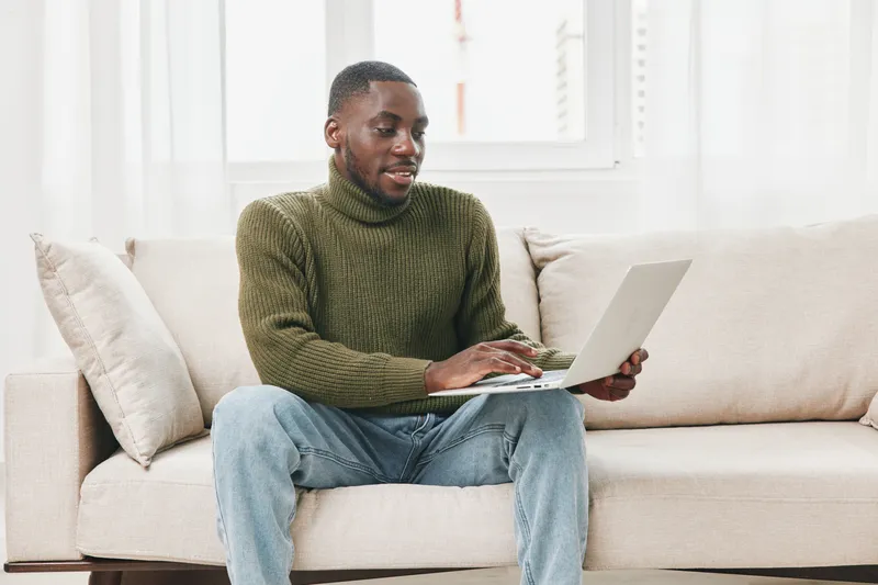 Young Black man sitting comfortably on a couch, using a laptop for work or study in a cozy indoor atmosphere