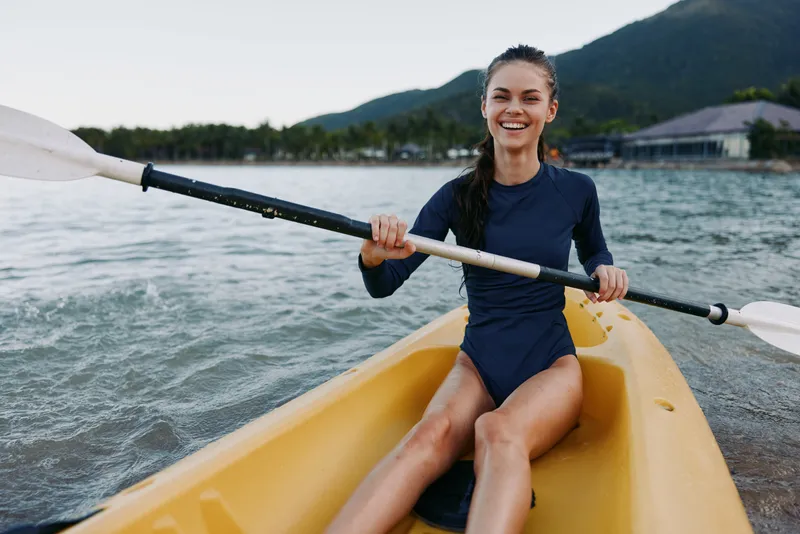 Smiling woman paddles a yellow kayak along calm water, gripping the paddle with confidence as she glides toward the shore, showcasing outdoor water sports and active relaxation.