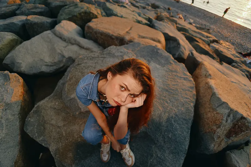 thoughtful woman sitting on large rocks near the beach during sunset, wearing denim and sneakers, gazing upward with a pensive expression