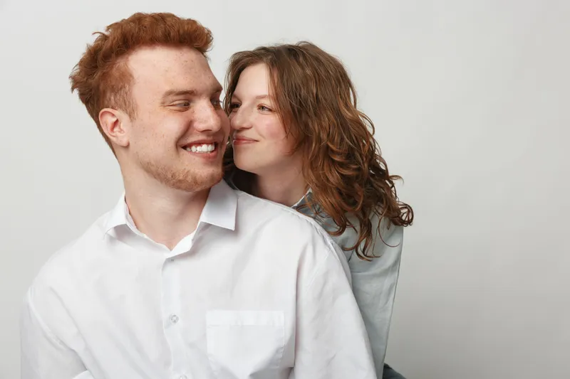 Happy young couple with red hair and curly hair smiling and embracing each other on a light background. Concept of love, relationship, affection, and togetherness.